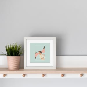 May include: A square white picture frame containing a collage print with a light blue background, a person in a white garment, and pink geometric shapes. Next to the frame is a small pink textured pot holding a green spiky artificial plant.