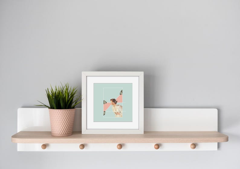 May include: A square white picture frame containing a collage print with a light blue background, a person in a white garment, and pink geometric shapes. Next to the frame is a small pink textured pot holding a green spiky artificial plant.