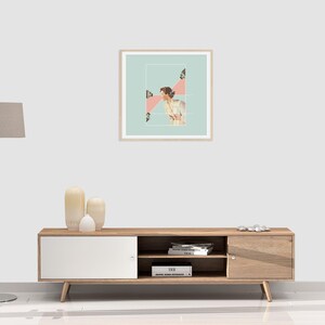 May include: A square framed wall art print featuring a collage of a person in a white shirt against a light blue background with pink geometric shapes and small camera icons, displayed in a light wood frame on a plain white wall.