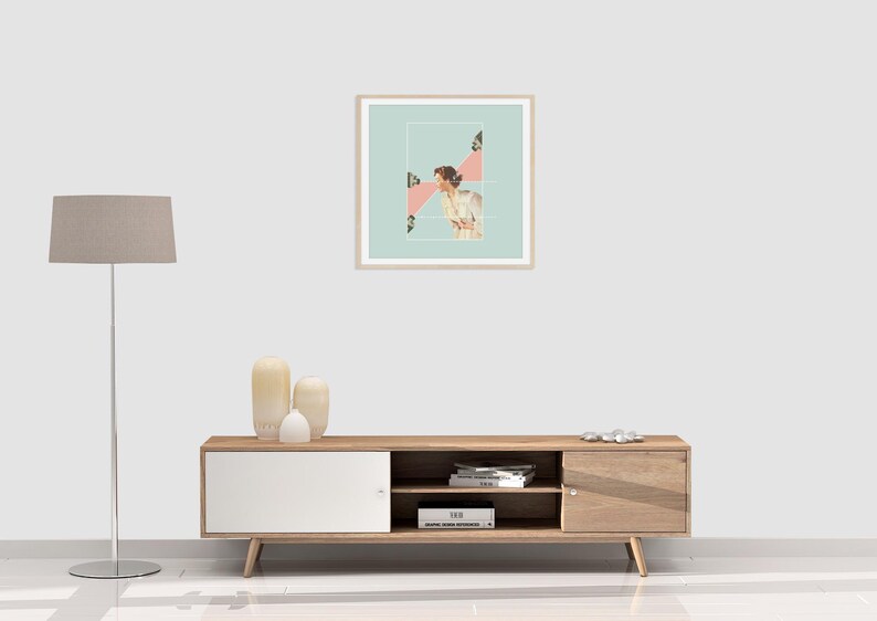 May include: A square framed wall art print featuring a collage of a person in a white shirt against a light blue background with pink geometric shapes and small camera icons, displayed in a light wood frame on a plain white wall.
