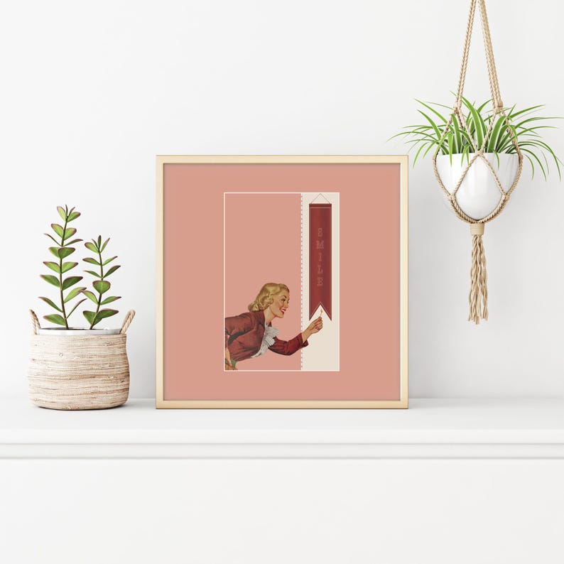 May include: A square framed print featuring a vintage-style illustration of a blonde person in a maroon jacket reaching toward a vertical maroon banner with the word SMILE printed in white. The artwork is set against a muted dusty rose background.