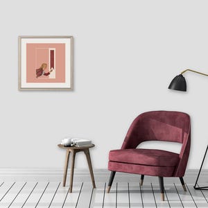 May include: A square framed art print featuring a person in a brown jacket reaching toward a vertical red rectangle against a dusty pink background, displayed on a wall above a dark red upholstered chair, a small wooden side table, and a floor lamp.