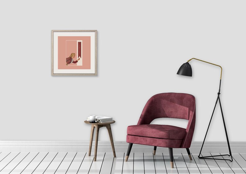 May include: A square framed art print featuring a person in a brown jacket reaching toward a vertical red rectangle against a dusty pink background, displayed on a wall above a dark red upholstered chair, a small wooden side table, and a floor lamp.