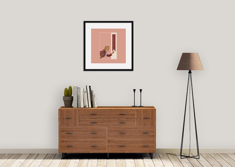 May include: A framed square art print featuring a person with blonde hair reaching toward a vertical red rectangle on a dusty pink background. The print is displayed above a wooden dresser with ten drawers, a potted cactus, books, and two black candlesticks.