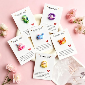 Op de afbeelding: Een verzameling "Pocket Hug" kaarten, elk met een kleine, kleurrijke zeedierbedel. De bedels omvatten een zeepaardje, een schelp met parel, een walvis, een octopus, een krab en een kwal. Elke kaart heeft een rood hart en tekst.