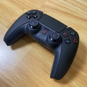 May include: A matte black wireless video game controller featuring a central PlayStation logo with red, yellow, green, and blue colors. The device includes two textured analog sticks, a directional pad, and four action buttons with colored symbols.