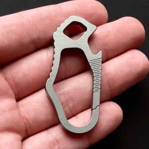 Puede incluir: Una pequeña multiherramienta plateada con un mosquetón. La herramienta cuenta con un abridor de botellas, un borde ranurado y un agarre texturizado. El diseño es compacto y parece estar hecho de metal.