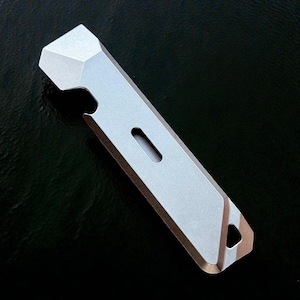May include: A silver metal multi-tool featuring a bottle opener, a small rectangular aperture, and a triangular hole at the end. The tool has a faceted top and a flat, rectangular body. The background is dark.