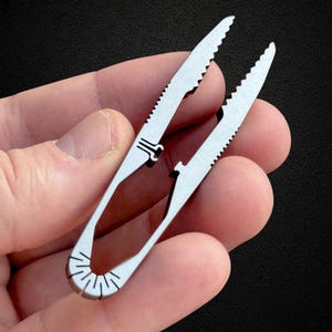 May include: A small, silver-colored multi-tool with serrated edges, held in a hand. The tool has a tweezer-like design with a curved handle and a series of lines etched into the base. The serrated edges suggest it may be used for cutting or gripping.