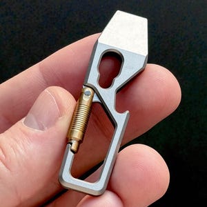 以下が含まれることがあります： キーホールデザイン、栓抜き、スプリング式留め金が付いた、シルバーとゴールドのマルチツール。コンパクトなサイズとメタリックな仕上がりを際立たせるように、手に持たれています。このツールは日常使いのために設計されています。