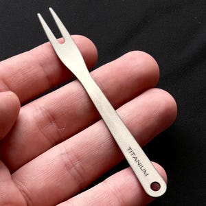 Peut inclure: Petite fourchette en titane argenté à deux dents. Le mot "TITANIUM" est imprimé sur le manche. Le manche comporte un petit trou à l'extrémité. La fourchette est tenue dans la main d'une personne.