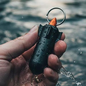 May include: A black, cylindrical keychain with an orange top, resembling a small bomb, is held in a hand with water droplets. The keychain has a silver ring attached. The background is a blurred view of water.
