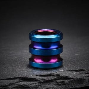 May include: A small, cylindrical bead with a layered design. The bead features a gradient of colours, including blue, purple, and pink. The bead is made of metal and has a polished finish. The bead is a decorative item.