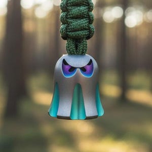 May include: A silver ghost-shaped pendant with teal accents and purple eyes, suspended from a green and black braided cord. The pendant has a unique design and is set against a blurred forest background.