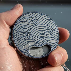 May include: A round bottle opener with a wave design in blue and gray. The opener is held in a hand with water droplets visible. The design features a stylized ocean wave pattern.