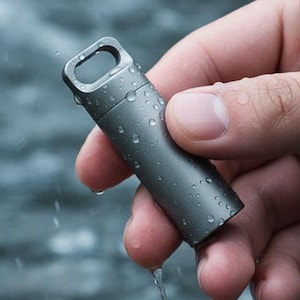 May include: A small, cylindrical, metallic gray container with a loop at the top. The container is held in a hand, with water droplets visible on its surface, suggesting it is waterproof. The background is a blurred view of water.