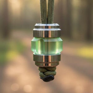 May include: A pendant with a green, translucent centre and silver-coloured metal accents. The pendant is suspended from a green cord. The background is a blurred view of a forest with sunlight.