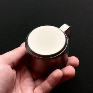 Pu&ograve; includere: Un piccolo contenitore rotondo in metallo spazzolato con una maniglia abbinata. Il coperchio &egrave; di una tonalit&agrave; d'argento pi&ugrave; chiara. Il contenitore &egrave; tenuto nella mano di una persona su uno sfondo scuro. Il design &egrave; minimalista e moderno.