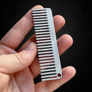 May include: A small, silver metal comb with closely spaced teeth. The comb has a rectangular shape with a rounded hole at one end. The comb is held in a hand against a dark background.