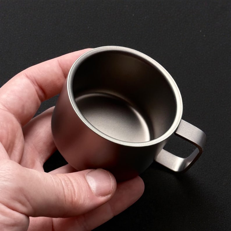 Pu&ograve; includere: Una piccola tazza in metallo spazzolato con un manico quadrato. La tazza &egrave; di un colore argento tenue e sembra essere in acciaio inossidabile. La tazza &egrave; tenuta in una mano su uno sfondo nero.