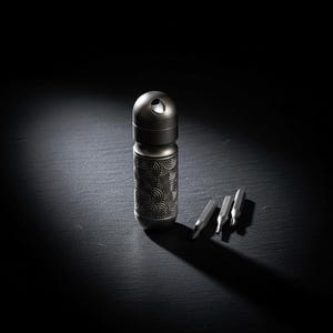 Może przedstawiać: Metalowe, szare narzędzie wielofunkcyjne z dekoracyjnym, okrągłym wzorem. Narzędzie ma cylindryczny korpus i zaokrąglony wierzch z otworem. Trzy srebrne bity są umieszczone obok narzędzia na ciemnej powierzchni.