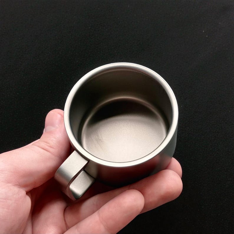 Pu&ograve; includere: Una piccola tazza in acciaio inossidabile con manico. La tazza &egrave; tenuta in mano, mostrando le sue dimensioni compatte e la finitura metallica. L'interno della tazza riflette la luce, evidenziando la sua superficie liscia. Il manico &egrave; rettangolare e attaccato al lato della tazza.