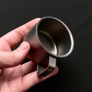 Pu&ograve; includere: Una piccola tazza in metallo spazzolato con un manico. La tazza &egrave; tenuta in mano su uno sfondo nero. La tazza ha un design semplice e minimalista, con un corpo cilindrico e un manico curvo.