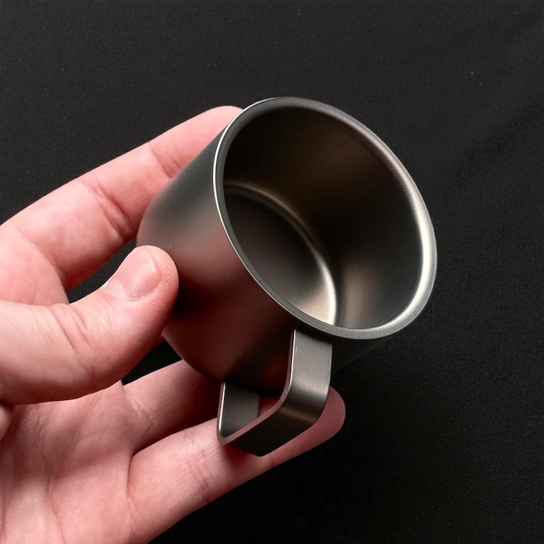 Pu&ograve; includere: Una piccola tazza in metallo spazzolato con un manico. La tazza &egrave; tenuta in mano su uno sfondo nero. La tazza ha un design semplice e minimalista, con un corpo cilindrico e un manico curvo.