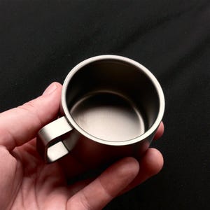 Pu&ograve; includere: Una piccola tazza in acciaio inossidabile con un manico curvo. La tazza ha un design semplice e minimalista e una finitura metallica lucida. La tazza &egrave; tenuta in mano su uno sfondo scuro. Ideale per il campeggio o i viaggi.