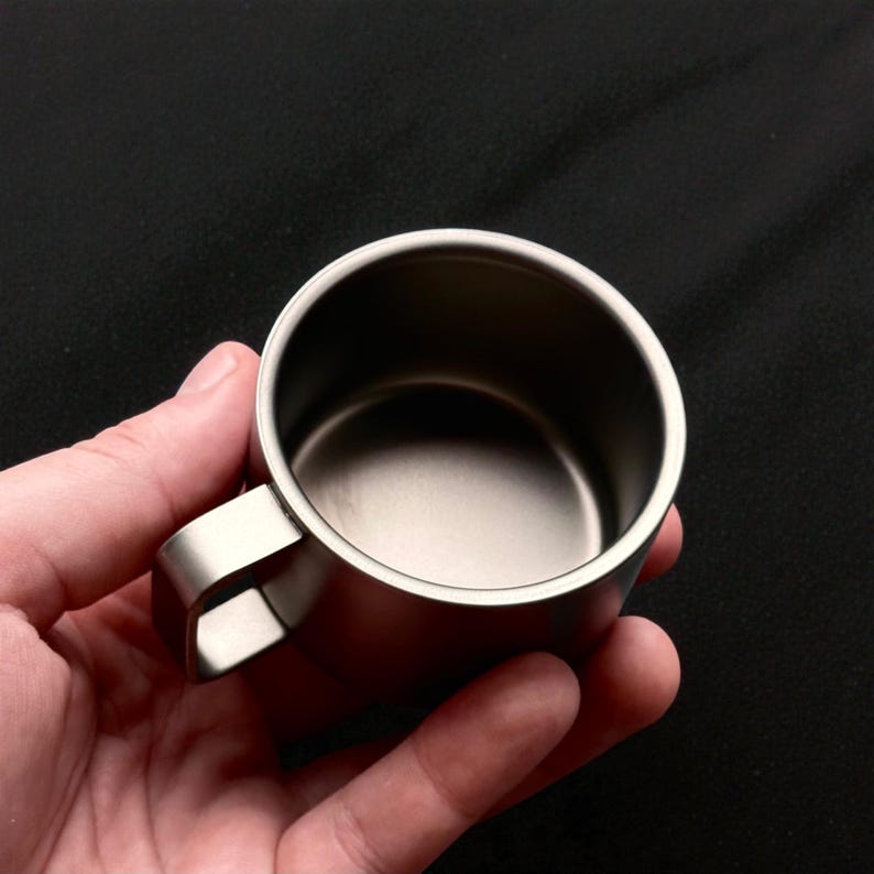 Pu&ograve; includere: Una piccola tazza in acciaio inossidabile con un manico curvo. La tazza ha un design semplice e minimalista e una finitura metallica lucida. La tazza &egrave; tenuta in mano su uno sfondo scuro. Ideale per il campeggio o i viaggi.