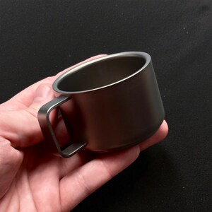 Pu&ograve; includere: Una piccola tazza grigio metallizzato con manico. La tazza &egrave; tenuta in mano su uno sfondo scuro. Il manico &egrave; un semplice anello rettangolare attaccato al lato. La tazza sembra essere fatta di un metallo resistente.