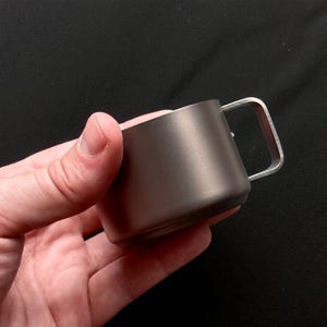 Pu&ograve; includere: Una piccola tazza di metallo grigio scuro con un manico argentato. La tazza &egrave; tenuta in mano su uno sfondo scuro. La tazza ha un design semplice e minimalista.