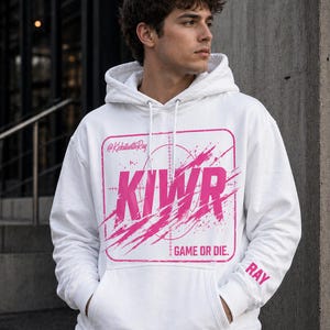 May include: White hoodie with a pink graphic that says "KIWR" inside a square with a target design and the words "GAME OR DIE." The sleeve has the name "RAY" printed on it. The hoodie has a front pocket and a hood.