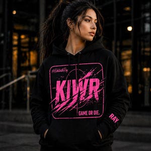 May include: Black hoodie with a pink graphic design. The design includes the letters "KIWR" inside a square with a target reticle and the words "GAME OR DIE." The sleeve has the name "RAY" printed on it. The hoodie has a drawstring hood and a front pocket.