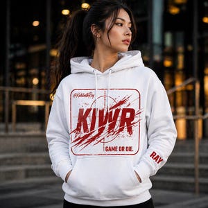 May include: White hoodie with a red graphic design. The design includes the letters "KIWR" inside a rectangular frame with a target and splatter effect. The text "GAME OR DIE." is below the graphic. The sleeve has the name "RAY".