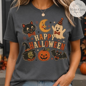 Könnte beinhalten: Ein dunkelgraues T-Shirt mit Rundhalsausschnitt und einem Halloween-Motiv im Vintage-Stil. Das Design zeigt eine schwarze Katze, eine Mondsichel, ein Gespenst mit Partyhut, eine Hexe, einen Kürbis und eine Fledermaus sowie den Text "HAPPY HALLOWEEN" in Orange.