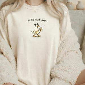 May include: Cream-colored t-shirt with the text "off to rope drop" above a cartoon goose wearing mouse ears and holding a coffee cup. The goose is walking with a determined expression. The shirt is worn by a person.