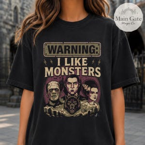 May include: A black short-sleeve t-shirt featuring a graphic print with the text WARNING: I LIKE MONSTERS. The design includes illustrations of Frankenstein's monster, a vampire, a werewolf, and the bride of Frankenstein above a graveyard scene.