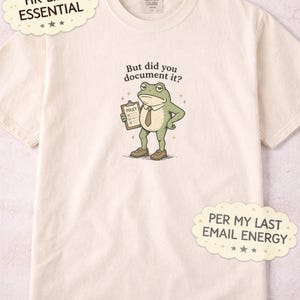 Op de afbeelding: Crèmekleurig T-shirt met een cartoonkikker met stropdas die een klembord met het woord "POLICY" vasthoudt. De tekst boven de kikker luidt "But did you document it?". Extra tekst bevat "HR LIFE ESSENTIAL" en "PER MY LAST EMAIL ENERGY".