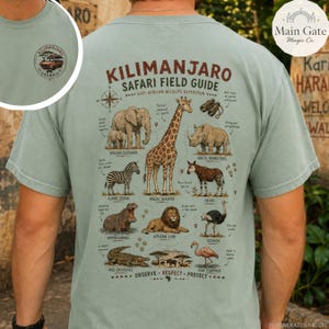 May include: A sage green t-shirt featuring a back graphic titled Kilimanjaro Safari Field Guide with illustrations of an elephant, giraffe, rhino, zebra, okapi, hippo, lion, ostrich, crocodile, and flamingo, plus a small front chest logo.