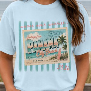 Könnte beinhalten: Hellblaues T-Shirt mit einem Vintage-Postkarten-Design. Die Postkarte zeigt "Greetings from Panama City Beach, Florida" mit einer Palme, einem Surfbrett und dem Text "Waves over worries". Die Zahl "2026" ist unten aufgedruckt.