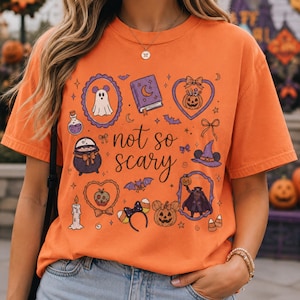 Könnte beinhalten: Orangefarbenes T-Shirt mit einem Halloween-Motiv. Das Design umfasst einen Geist, ein Buch, einen Kürbis, einen Hexenhut und die Worte "not so scary". Das T-Shirt hat kurze Ärmel und einen Rundhalsausschnitt.