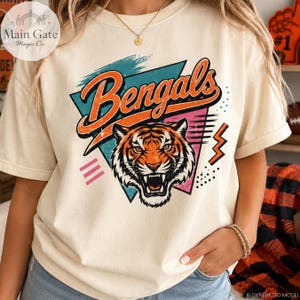 Może przedstawiać: Kremowy t-shirt z grafiką ryczącej głowy tygrysa umieszczoną centralnie na tle turkusowo-różowego trójkąta. Słowo Bengals jest nadrukowane dużą pomarańczową kursywą nad tygrysem. Wzór zawiera różowe linie oraz kropki jako elementy dekoracyjne.
