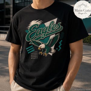 Può includere: Una t-shirt nera a maniche corte con una stampa grafica di un'aquila calva in volo con ali verdi e bianche. La parola Eagles è scritta in un grande carattere corsivo verde sopra l'aquila. Forme geometriche circondano il design centrale.
