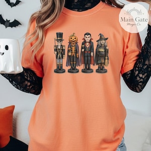 May include: An orange short-sleeved t-shirt featuring a graphic print of four Halloween-themed figures: a skeleton nutcracker, a pumpkin-headed character, a vampire, and a witch. The shirt is layered over a black long-sleeved lace top.