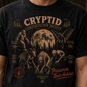 Puede incluir: Camiseta negra con un diseño de la Cryptid Conservation Society. El gráfico incluye ilustraciones de Bigfoot, Mothman, Jersey Devil, Skinwalkers y Fresno Nightcrawler, con el texto "Preserve Their Habitat."