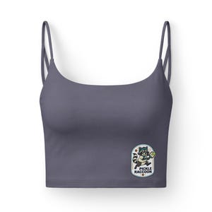 Op de afbeelding: Een leisteen grijs crop top met dunne spaghettibandjes. De top heeft een kleurrijke patch met een cartoon wasbeer die pickleball speelt, met de tekst "PICKLE RACCOON".