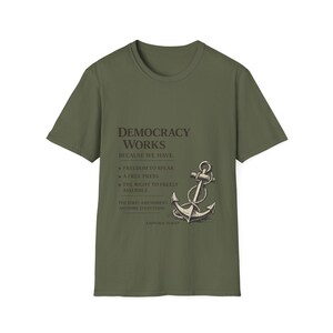 Puede incluir: Camiseta verde oliva con el texto "DEMOCRACY WORKS" y un gráfico de ancla. La camiseta enumera libertades como la de expresión y prensa. La frase "THE FIRST AMENDMENT ANCHORS EVERYTHING" también está impresa.
