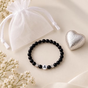 Könnte beinhalten: Ein Perlenarmband mit glatten, schwarzen, runden Perlen, zwei kleinen silberfarbenen Sternanhängern und einer einzelnen weißen runden Perle mit dem aufgedruckten schwarzen Buchstaben A, dazu ein weißer, transparenter Beutel und ein silbernes Herz.