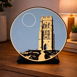 May include: A round, illuminated art piece featuring a stylized illustration of a tower on a hill. The artwork is set against a light blue background with a white circle. The piece is mounted on a black stand.